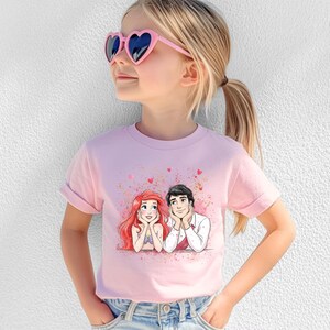 A young girl wearing a pink t-shirt with a cartoon image of two characters, one with red hair and the other with blonde hair, smiling and looking up at each other.