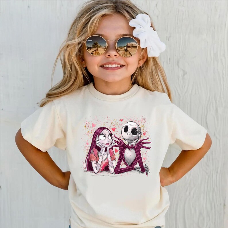 A young girl wearing a white t-shirt with a graphic of two cartoon characters, one of which is Jack Skellington, smiling and looking at the other character.