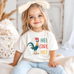 A young girl with blonde hair wearing a white t-shirt with a colorful rooster graphic and the text "HEI GIRL HEI" is sitting on a bed.