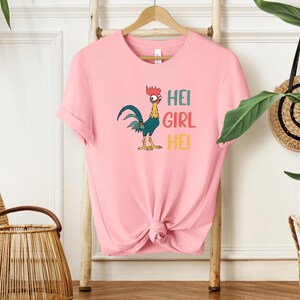 A pink t-shirt with a cartoon rooster and the text "HEI GIRL HEI" printed on it, hanging on a wooden hanger.