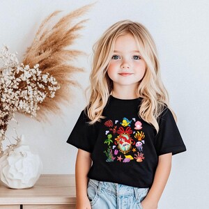 A young girl with blonde hair and blue eyes is smiling and posing in front of a white wall with a vase of white flowers and a wooden table.