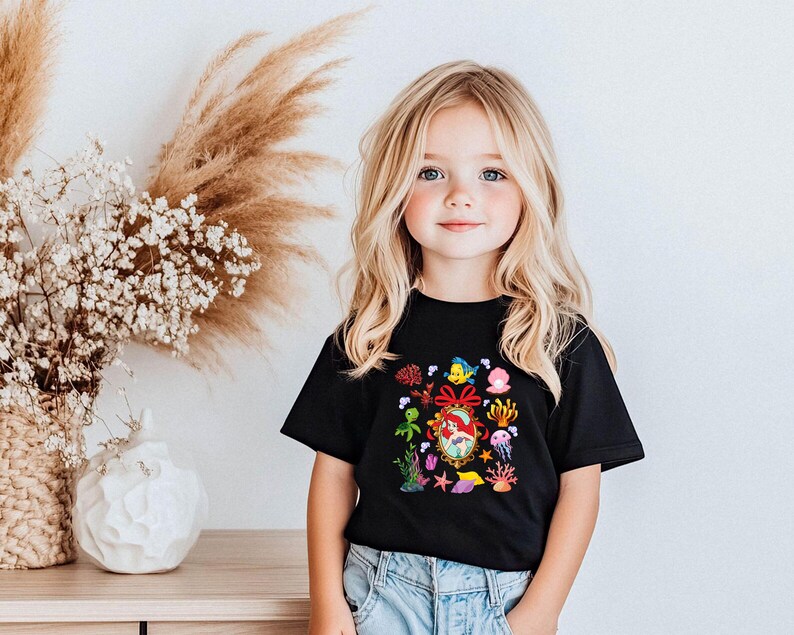 A young girl with blonde hair and blue eyes is smiling and posing in front of a white wall with a vase of white flowers and a wooden table.