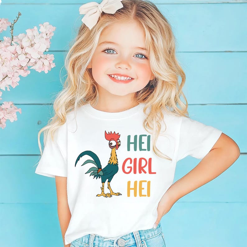 A young girl with blonde hair is smiling and wearing a white t-shirt with a graphic of a rooster and the text "HEI GIRL HEI" printed on it.