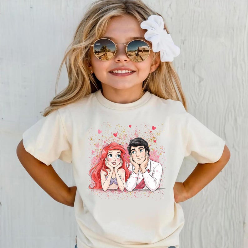 A young girl wearing a white t-shirt with a graphic of two cartoon characters, a man and a woman, smiling and looking at each other.