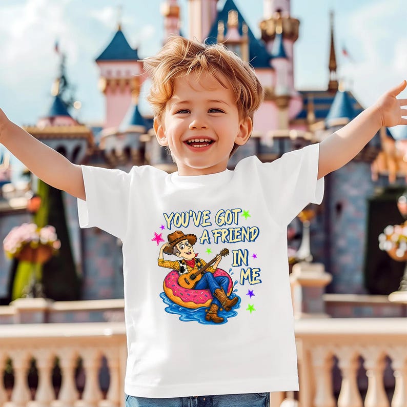 A young boy with blond hair is smiling and raising his arms, wearing a white t-shirt with a cartoon character and text printed on it.