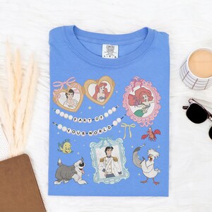 a blue t-shirt with a Disney-themed design, including characters from the animated film "The Little Mermaid". The t-shirt is laid out on a white surface, accompanied by a mug, sunglasses, and a notebook.