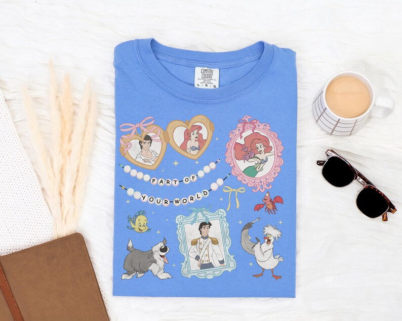 a blue t-shirt with a Disney-themed design, including characters from the animated film "The Little Mermaid". The t-shirt is laid out on a white surface, accompanied by a mug, sunglasses, and a notebook.