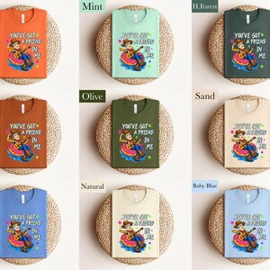 The image is a collage of nine different t-shirt designs, each with a unique color and design. The t-shirts are arranged in a 3x3 grid, with each row containing three shirts. The designs include a character, a message, and a color. The t-shirts are displayed on a woven basket, which adds a rustic touch to the overall presentation.