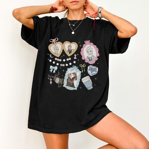 A person wearing a black t-shirt with various cartoon-style illustrations and text, including characters and objects such as hearts, a cup, and a mirror.