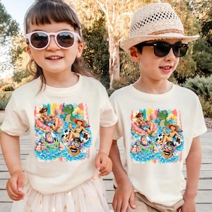 May include: Two off-white short-sleeve t-shirts featuring a colorful graphic print of various animated characters in a swimming pool. The design includes figures wearing sunglasses and swim gear, floating on inflatable rings in blue water.