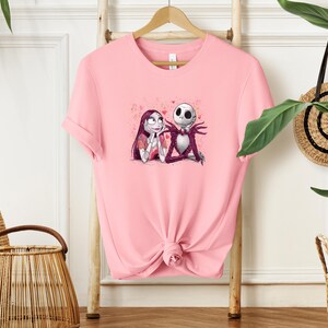 A pink t-shirt with a graphic of two cartoon characters, Jack Skellington and Sally, is hanging on a wooden hanger.