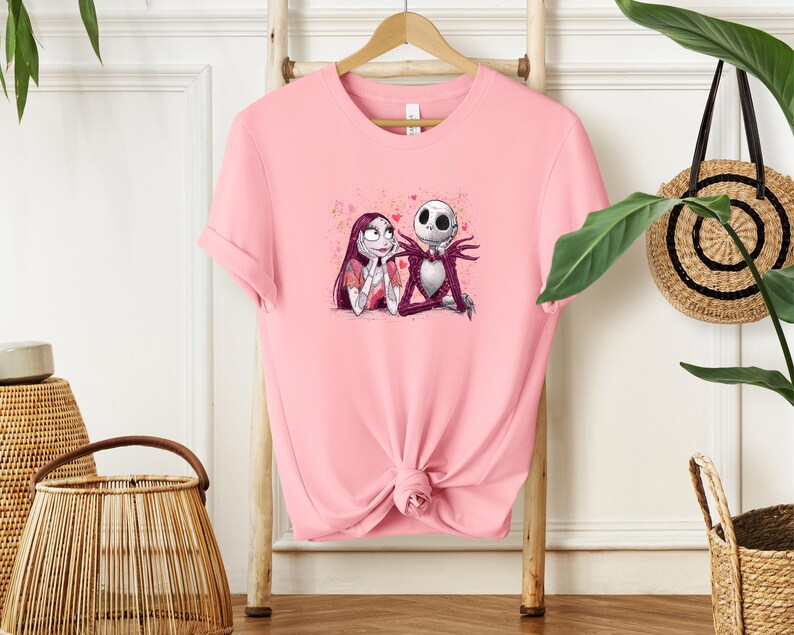 A pink t-shirt with a graphic of two cartoon characters, Jack Skellington and Sally, is hanging on a wooden hanger.