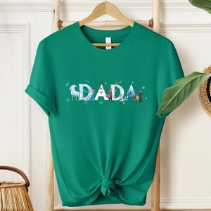 A green t-shirt with the word "DADA" printed on it, hanging on a wooden hanger.