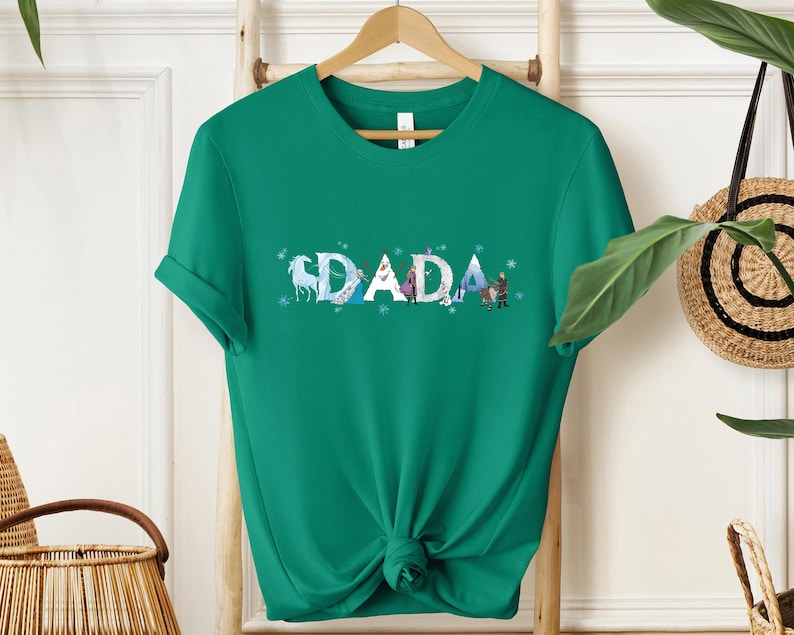 A green t-shirt with the word "DADA" printed on it, hanging on a wooden hanger.