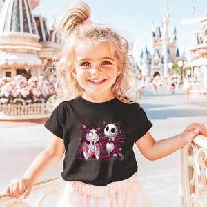 A young girl with blonde hair is smiling and posing in front of a Disney theme park, wearing a black t-shirt with a graphic of Jack Skellington and Mary Jane.
