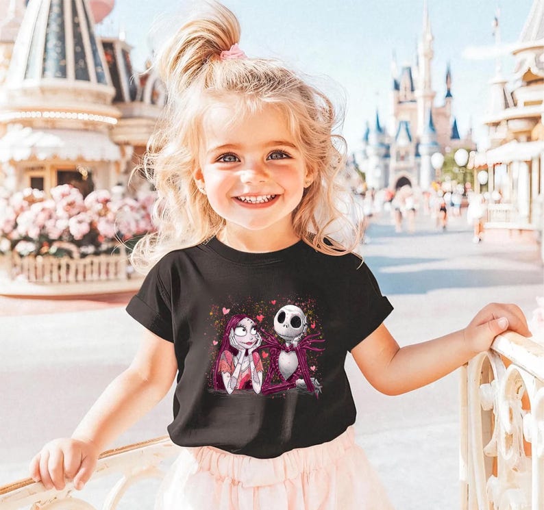 A young girl with blonde hair is smiling and posing in front of a Disney theme park, wearing a black t-shirt with a graphic of Jack Skellington and Mary Jane.