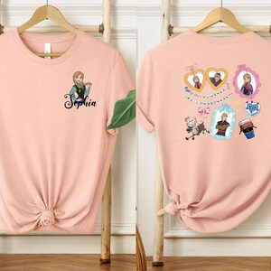 two pink t-shirts hanging on wooden hangers. The t-shirt on the left has a name tag with the name "Sophia" printed on it, while the t-shirt on the right has various cartoon-style illustrations and text on it.