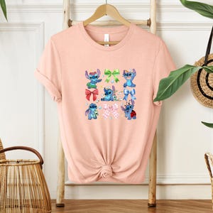 A pink t-shirt with a graphic design of various cartoon characters, including Stitch, is displayed on a wooden hanger.