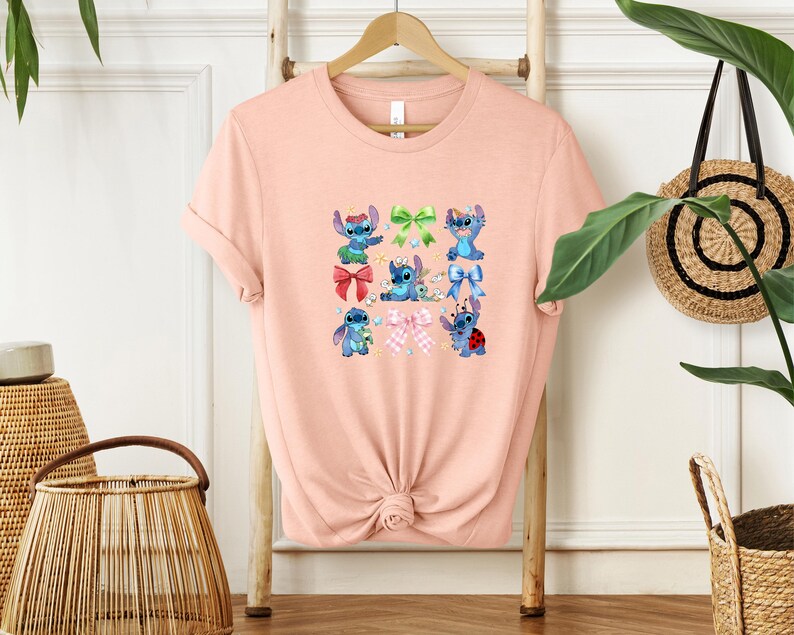 A pink t-shirt with a graphic design of various cartoon characters, including Stitch, is displayed on a wooden hanger.