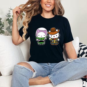 A woman sitting on a couch wearing a black t-shirt with a graphic of two cartoon characters, one wearing a green outfit and the other a cowboy hat.