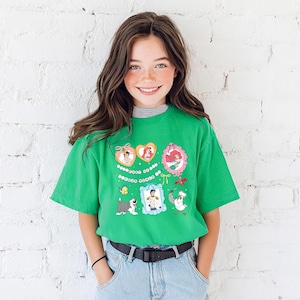 A young girl with long brown hair is smiling and wearing a green t-shirt with colorful cartoon characters on it, standing in front of a white brick wall.