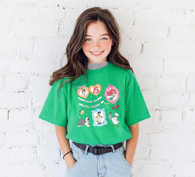 A young girl with long brown hair is smiling and wearing a green t-shirt with colorful cartoon characters on it, standing in front of a white brick wall.