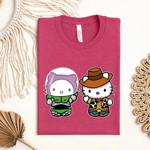 a pink t-shirt with a graphic of two cartoon characters, a cat and a character wearing a cowboy hat, along with a tag. The t-shirt is laid out on a white surface, surrounded by various decorative items such as a macrame wall hanging, a woven mat, and a leaf.