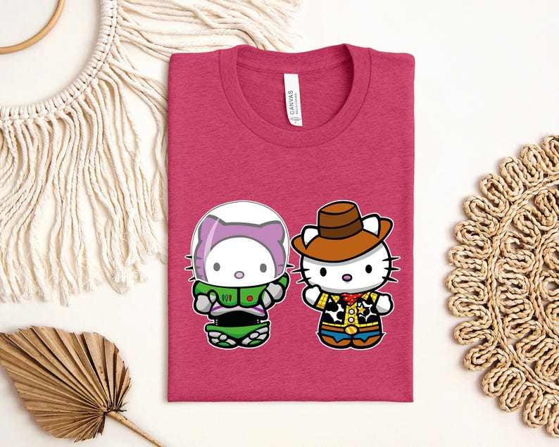 a pink t-shirt with a graphic of two cartoon characters, a cat and a character wearing a cowboy hat, along with a tag. The t-shirt is laid out on a white surface, surrounded by various decorative items such as a macrame wall hanging, a woven mat, and a leaf.
