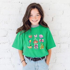 A young girl with long brown hair is smiling and posing in front of a white brick wall. She is wearing a green t-shirt with a colorful graphic design featuring various cartoon-like characters and elements.