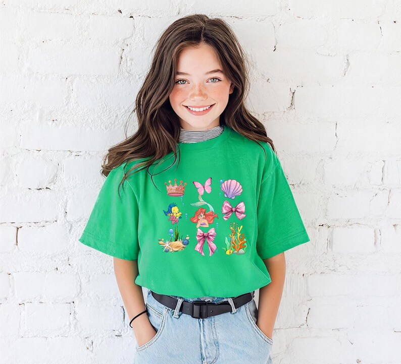 A young girl with long brown hair is smiling and posing in front of a white brick wall. She is wearing a green t-shirt with a colorful graphic design featuring various cartoon-like characters and elements.