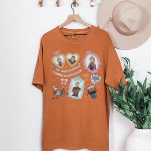 a brown t-shirt hanging on a wooden hanger, adorned with various cartoon-style illustrations and characters. The t-shirt is displayed against a white wall, with a hat and a potted plant visible in the background.