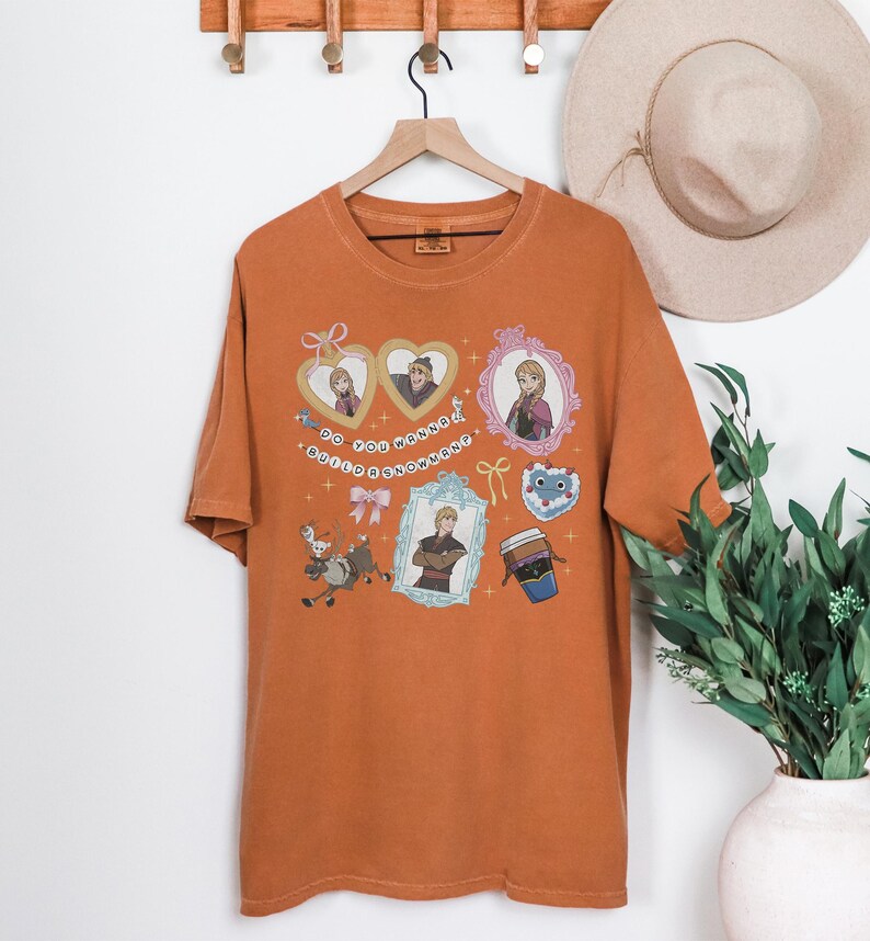 a brown t-shirt hanging on a wooden hanger, adorned with various cartoon-style illustrations and characters. The t-shirt is displayed against a white wall, with a hat and a potted plant visible in the background.