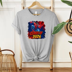 A gray t-shirt with a graphic design featuring a superhero character and the text "CLASS OF 2026" printed on it, hanging on a wooden hanger.