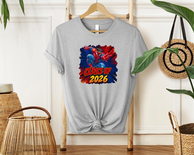 A gray t-shirt with a graphic design featuring a superhero character and the text "CLASS OF 2026" printed on it, hanging on a wooden hanger.