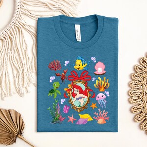 a blue t-shirt with a colorful design of various sea creatures and elements, including a mermaid, sea turtle, jellyfish, coral, and seaweed.