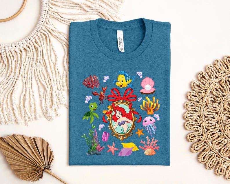 a blue t-shirt with a colorful design of various sea creatures and elements, including a mermaid, sea turtle, jellyfish, coral, and seaweed.
