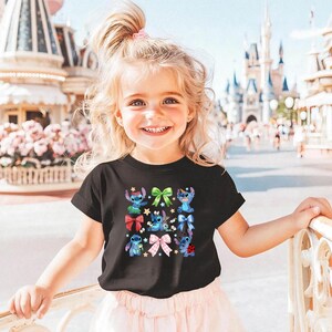 A young girl with blonde hair is smiling and posing in front of a Disney theme park, wearing a black t-shirt with a colorful design featuring characters from the Disney movie "Stitch".