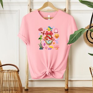 A pink t-shirt with a colorful graphic design of various sea creatures and plants, hanging on a wooden hanger.