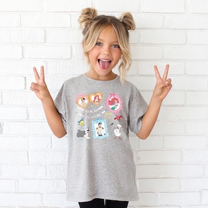 A young girl with blonde hair is making a peace sign with her hands, wearing a gray t-shirt with a cartoon character design.