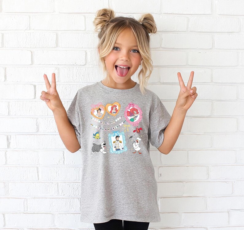 A young girl with blonde hair is making a peace sign with her hands, wearing a gray t-shirt with a cartoon character design.