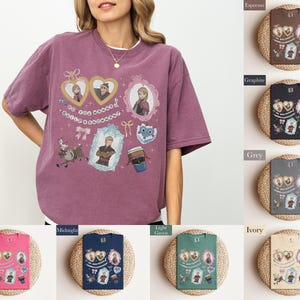 A woman wearing a purple t-shirt with various cartoon characters and designs, including a heart and a cup, is standing next to a display of different colored books or notebooks.