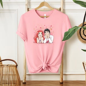 A pink t-shirt with a cartoon illustration of two people, likely a couple, is hanging on a wooden hanger. The t-shirt is displayed on a wooden stand, and there are some plants and baskets in the background.