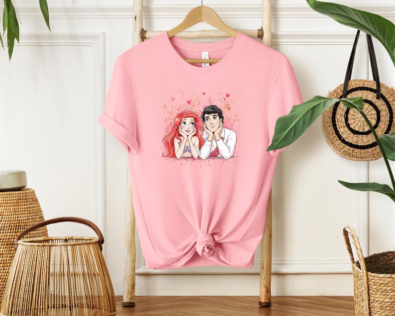 A pink t-shirt with a cartoon illustration of two people, likely a couple, is hanging on a wooden hanger. The t-shirt is displayed on a wooden stand, and there are some plants and baskets in the background.