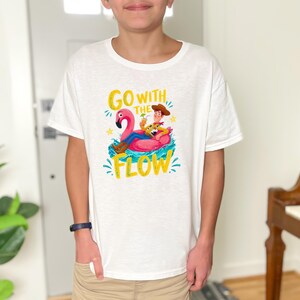 Può includere: Una t-shirt bianca a maniche corte con la grafica di un personaggio cowboy seduto su un salvagente a forma di fenicottero rosa. Il testo "GO WITH THE FLOW" è stampato in giallo attorno al personaggio, circondato da spruzzi d'acqua blu.