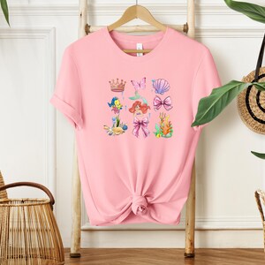 A pink t-shirt with a mermaid design and various sea-themed illustrations is displayed on a wooden hanger.