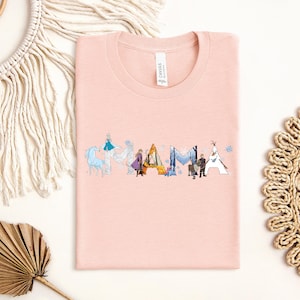a pink t-shirt with a graphic design of various characters from the Disney movie Frozen, including Elsa, Anna, Olaf, and other popular characters. The t-shirt is laid out on a white background, surrounded by other items such as a macrame decoration, a woven mat, and a leaf.