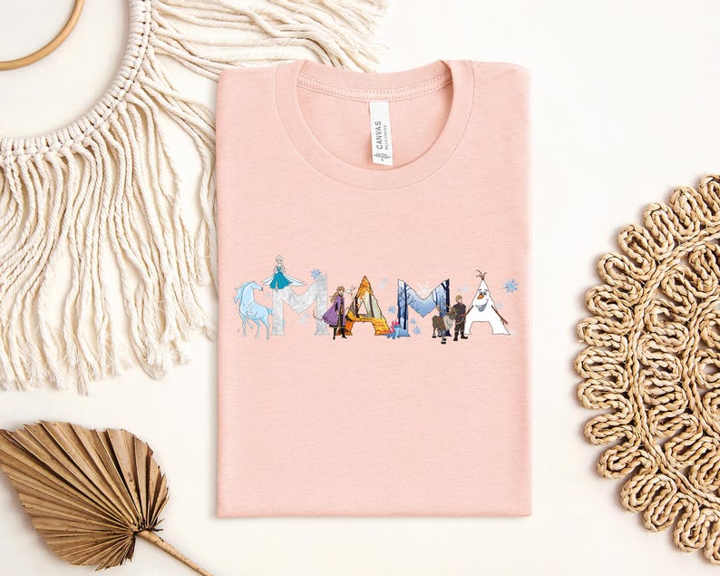 a pink t-shirt with a graphic design of various characters from the Disney movie Frozen, including Elsa, Anna, Olaf, and other popular characters. The t-shirt is laid out on a white background, surrounded by other items such as a macrame decoration, a woven mat, and a leaf.