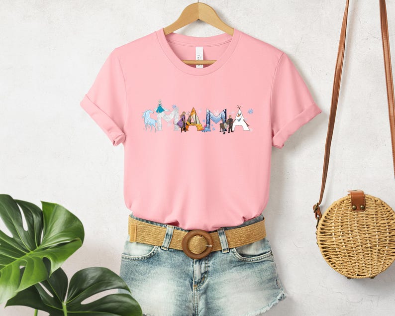 A pink t-shirt with a graphic design of various cartoon characters, including a horse, a dog, and a person, is displayed on a hanger. The t-shirt is paired with denim shorts and a round woven purse, creating a casual and stylish outfit.