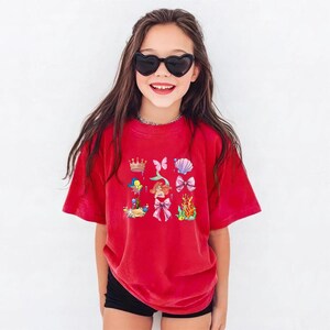 A young girl wearing a red t-shirt with a mermaid design and sunglasses, smiling at the camera.