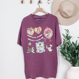 A purple t-shirt with a Disney-themed design hanging on a wooden hanger, along with a beige hat and a potted plant.